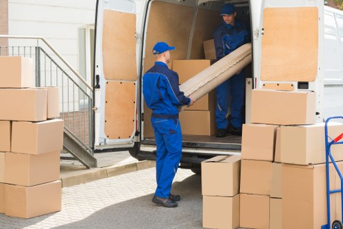 Two removal workers dressed in blue uniforms are loading cardboard boxes into a white removals van parked on a residential street near a house in Brixton. The van's rear doors are open, revealing a wooden panel interior. One worker is holding a large rolled-up cardboard tube, while the other is passing or receiving it from inside the van. Several stacked cardboard boxes are positioned on the ground nearby, some directly in front of the van and others piled to the side, suggesting a house move or relocation process typical in Brixton. The scene is outdoors, with natural daylight illuminating the workspace, and a metal staircase and residential building visible in the background, emphasizing the local street environment. The scene depicts a professional removals service focused on efficient packing and loading, with emphasis on household furniture and personal items being prepared for transport in a familiar London neighbourhood.