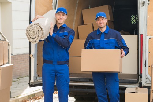 Two professional movers dressed in blue uniforms and matching caps are standing outside a van used for house removals, with the rear doors open. The mover on the left is smiling and carrying a rolled-up rug or blanket over his shoulder, while the one on the right is holding a medium-sized cardboard box. The interior of the van reveals a variety of packed cardboard boxes of different sizes stacked in the background, ready for transportation. The scene takes place on a residential driveway or street in Brixton, with part of a house and some garden features visible nearby. The environment looks clean and organized, with natural daylight illuminating the scene, highlighting the textures of the cardboard boxes, the fabric of the uniforms, and the wooden and metallic details of the boxes and van. The image reflects professional house removal services, emphasizing the careful handling of household items and the efficient teamwork involved in moving belongings locally within London postcode areas near Brixton.