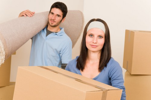 A young man and woman with light skin and brown hair in a residential interior, likely a living room or hallway, surrounded by unpacked cardboard boxes of various sizes, some sealed with packing tape, indicating a house move. The woman in the foreground is holding a medium-sized box, wearing a white headband and a blue top, with a neutral expression. The man in the background is standing behind her, carrying a large, light-colored piece of furniture, possibly a sofa or wardrobe, over his shoulder, dressed in a light blue shirt. The scene is well-lit, with neutral-toned walls and a simple, tidy environment, suggesting they are in the process of relocating. The activity relates to house removals or local moving services provided by Brixton Man with Van, based in the Brixton area. This image captures the moments of packing and lifting items typical of house clearance or apartment moves within London, especially around the postcode area associated with Brixton.