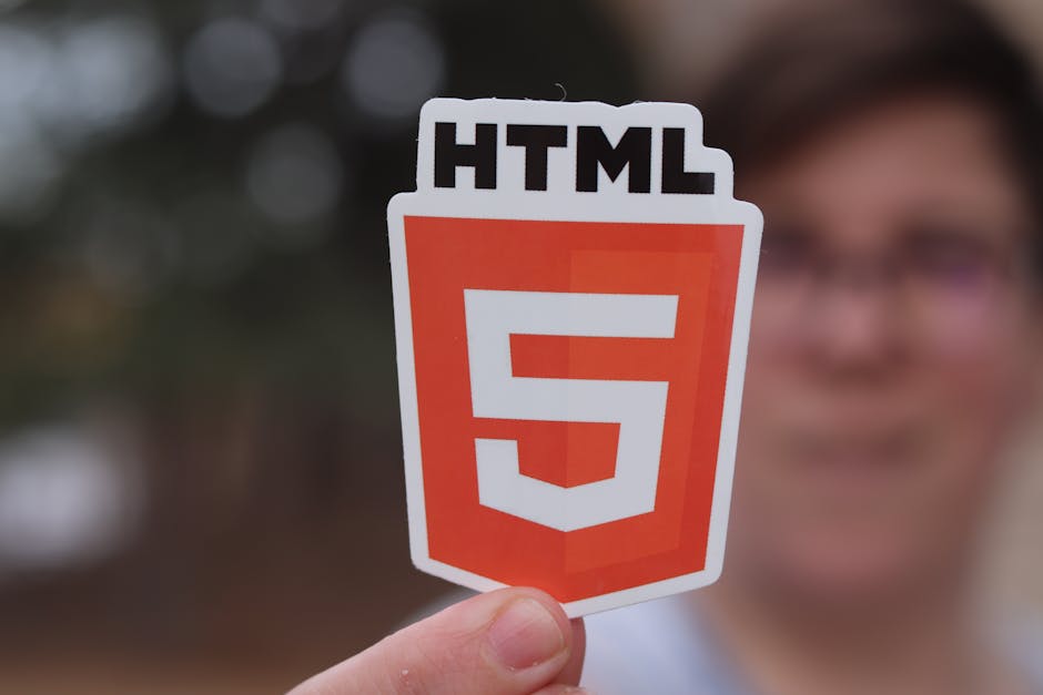 A person holding a sticker with the HTML5 logo, featuring a white and orange shield shape with the text 'HTML' above the number '5', in front of a blurred outdoor background. The sticker is being held