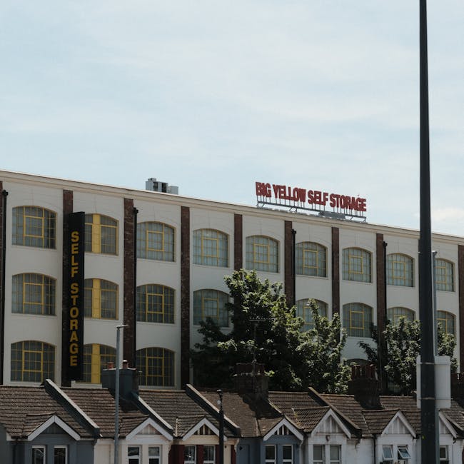 A close-up view of a multi-storey residential building with pink exterior walls, featuring several balconies with metal railings, some of which have glass panels, and windows adorned with curtains. On
