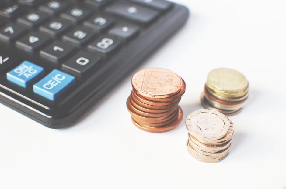 Close-up image of a black calculator with blue function keys, positioned next to three neatly stacked piles of British coins in various denominations, including pennies and pounds, on a white surface.