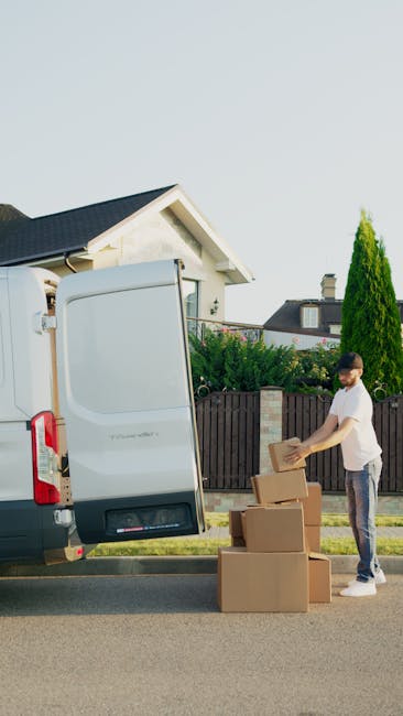 A man wearing a blue jacket, black vest, black pants, white sneakers, and a grey cap is pushing a hand truck loaded with two large cardboard boxes outside a grey commercial van with its rear doors ope